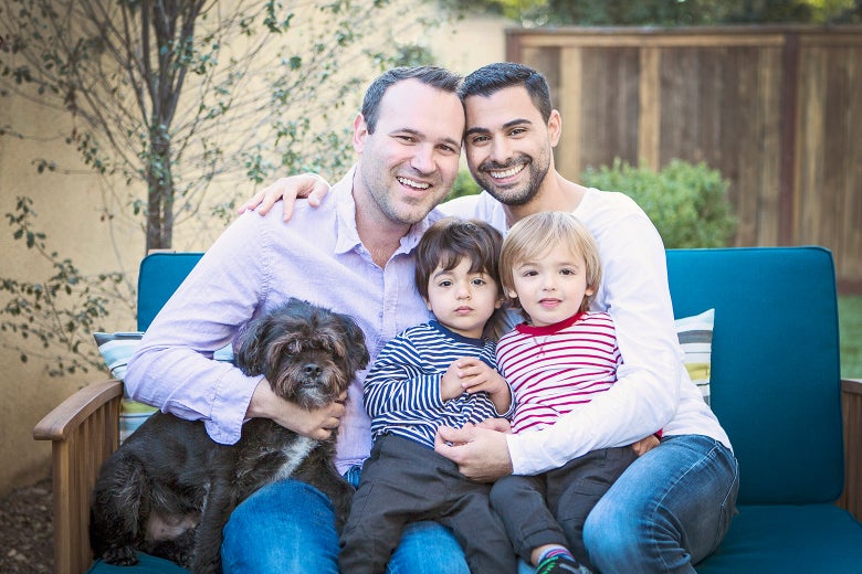A couple holds their two toddler sons in their lap with a dog at their side.